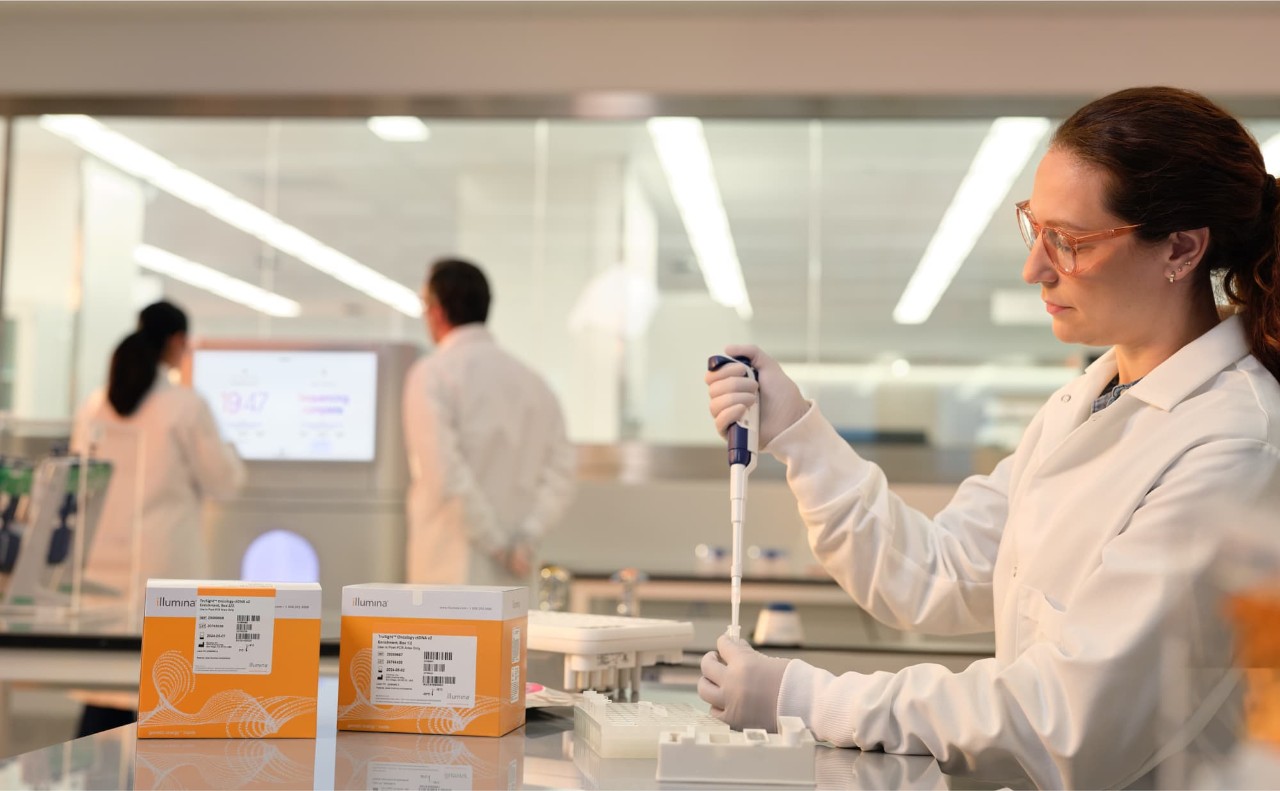 Profile image of a female scientist using a single pipette into a tube, two boxes of TruSight Oncology ctDNA v2 Enrichment library prep boxes on the lab bench with other NovaSeq X consumables; two scientists and a NovaSeq X instrument are blurry in the background.